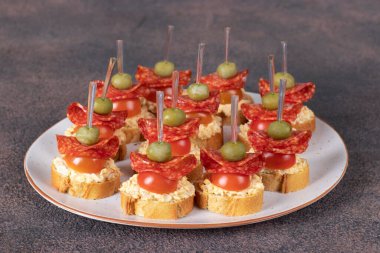 Sosisli salamlı kanepeler, vişneli domatesler ve kahverengi arka planda beyaz tabakta yeşil zeytinler.