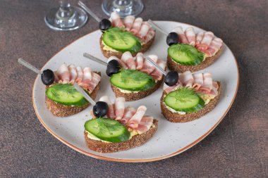 Domuz pastırmalı sandviç, salatalık ve siyah zeytinli tost beyaz tabakta çavdar ekmeğinden.