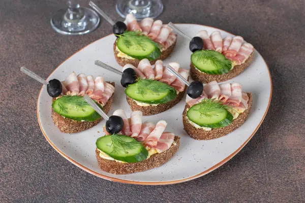 Domuz pastırmalı sandviç, salatalık ve siyah zeytinli tost beyaz tabakta çavdar ekmeğinden.