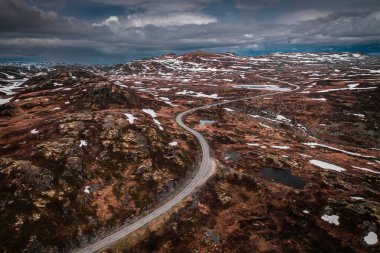 Lonesome road through the landscape of Hardangervidda National Park in Norway, snow fields and dark clouds in the sky, from above