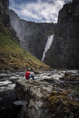 Man sitting on rock watching at Voringsfossen waterfall with steep mountain cliffs in the valley at Hardangervidda National Park in Norway