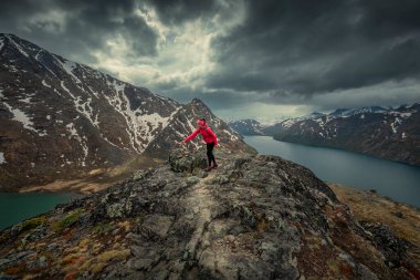 Woman building stone pyramid during hike along blue lakes in mountain landscape of Knutshoe in Jotunheimen National Park in Norway, mountains of Besseggen in background, dramatic cloudy sky