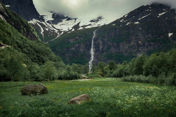 Waterfall with meadows in the mountains of Briksdalsbreen glacier in Jostedalsbreen national park in Norway, moody atmosphere with clouds in sky, rocks in green grass
