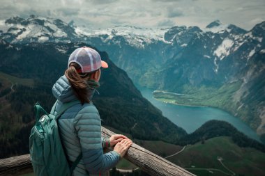 Female hiker with backpack looking at turquoise lake Knigssee from above viewpoint of mountain peak Jenner at Berchtesgaden Bavaria, snow-capped mountains in the background