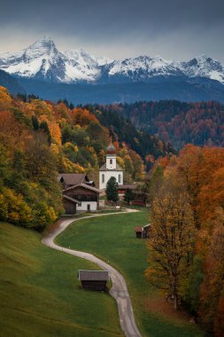 Bavarian Alps with church of Wamberg in Garmisch-Partenkirchen during autumn, snow-covered mountains in the background, dramatic cloudy sky, colored leaves and trees and huts and road in foreground, Bavaria Germany