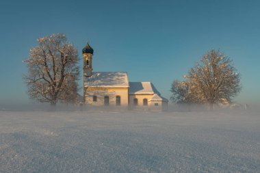 Bavarian church of Raisting with trees and snow and mist during winter and sunset, snow field in the foreground, blue sky day, Bavaria Germany