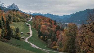 Timelapse of Bavarian Alps with church of Wamberg in Garmisch-Partenkirchen during autumn, snow-covered mountains in the background, moving clouds in sky, colored leaves and trees and huts and road in foreground, Bavaria Germany