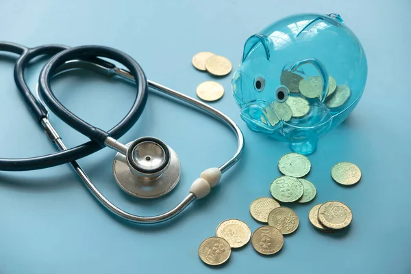 Concept of saving for Medical insurance costs or expense and financial checkup. Transparent piggy bank, coins and stethoscope on blue background.