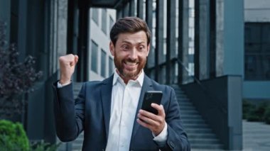 Heyecanlı, şaşırmış bir adam açık havada telefon ekranına bakıyor zaferi kutlayan iyi haberleri okuyor hevesli iş adamı başarısından dolayı seviniyor akıllı telefondan başarılı bir mesaj alıyor.