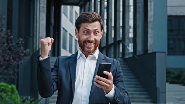 兴奋而惊讶的男人站在户外看手机屏幕读着好消息庆祝胜利热情洋溢的商人成功人士在智能手机上收到了成功的信息 — 图库视频影像