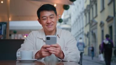Gülümseyen Asyalı turist elinde cep telefonu ile oturmuş internetten bilet ayırtıyor. Sosyal ağdaki arkadaşlarıyla ücretsiz internet üzerinden iletişim kuruyor.