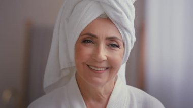Mutlu gülümseyen, çekici, yaşlı, olgun, yaşlı, bornoz giyen, beyaz havlu takan 60 'lı yaşlarda yaşlı bir kadın. Yüzünde buruşuk bir cilt bakımı prosedürüyle kameraya bakıyor.