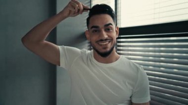 Gülümseyen Latin Hintli sakallı adam kameraya bakıyor saç fırçasıyla saçlarını tarıyor saçlarını düzeltiyor gülüşü sağlıklı, güzel saç stili, rutin saç kesimiyle mutlu.