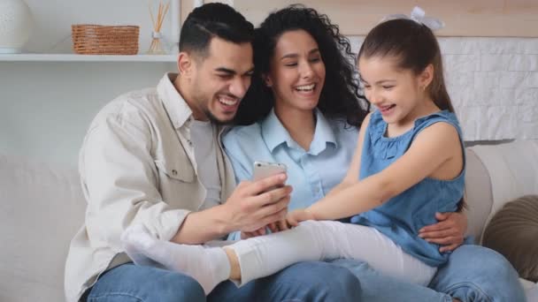 多种族 多族裔 快乐的家庭西班牙裔阿拉伯白人父母带着小女儿坐在沙发上看搞笑视频电影动画片 用手机视频叫笑得很开心 — 图库视频影像