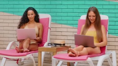 Bikini giyen iki seksi bayan spa tatil beldesi Kafkasyalı ve İspanyol kadın blogcularının plajlarında dizüstü bilgisayarlarıyla serbest çalışıyorlar. El hareketi yaparak yaz tatili için internet üzerinden seyahat rezervasyonu yapmalarını öneriyorlar.