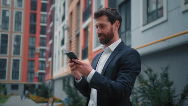 ハンサムな成功したビジネスマンの白人男性は 途中で検索情報をオンラインテキストメッセージ遠くのチャットで屋外の使用の携帯電話のデジタルモバイルアプリを動作市内の近代的な住宅ビルの近くに立つ — ストック動画