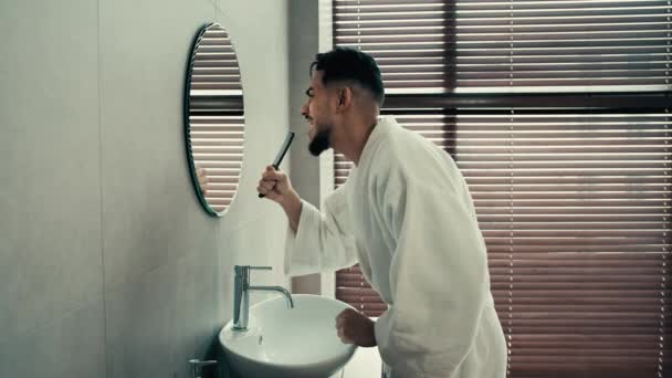 侧面看幽默滑稽无忧无虑的拉美裔拉美裔印度人和镜中人跳舞嬉戏身穿白色浴衣的男人站在浴室里唱歌早上在刷子里玩乐 — 图库视频影像
