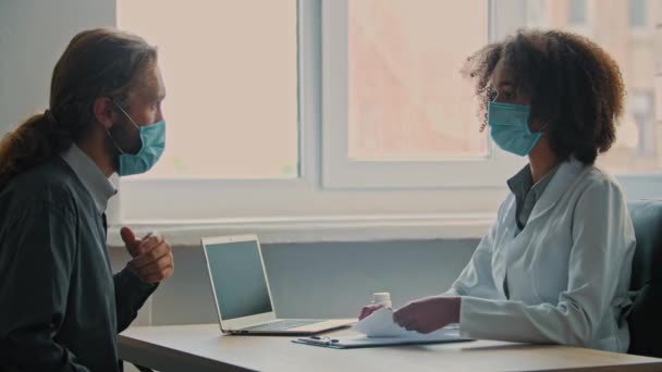 専門医アフリカの女性セラピストは病気の男性に相談します患者はマスクを身に着けています病気は病院で医療処方を必要とします女性看護師は薬治療薬局のコンセプトでボトルを与えます — ストック動画