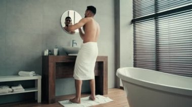Uzun menzilli bir manzara, ellerinde beyaz banyo havlusuyla dans eden komik bir adam, kalçalarına sarılmış aynaya bakıyor, pohpohlanarak kazanıyor, duştan sonra dans ederek müzik eşliğinde eğleniyor.