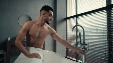 Milenyum seksi, çıplak, kaslı Arap adam, sakallı, havluyla kalçasına sarılmış banyo küvetinde oturmuş musluğu değiştiriyor, sıcak suyu tadıyor ve banyo yapmaya hazırlanıyor.