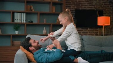 Kafkasyalı erkek bebek bakıcısı baba kanepeye uzanmış, sevgi dolu kız çocuğuyla oynuyor, çocukları gıdıklıyor, oyun oynuyor, birlikte eğleniyor tatilde neşeli bir çocukluk geçiriyor, mutlu bir aileye gülüyor.