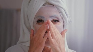 Banyo yapan yaşlı, olgun, beyaz bir kadının portresi. Kafasına havlu sar. Büyükanne, kadın yüz bakımı güzellik kozmetik prosedürüne cilt maskesi sür.