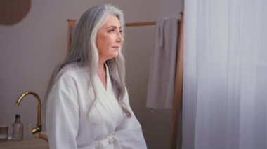 Düşünceli, yaşlı, 60 'lı yaşlarda, beyaz bornozlu, 50' li yaşlarda, beyaz, yaşlı, yaşlı, beyaz bir kadın. Evdeki tuvalette duruyor. Sakin, gri saçlı büyükanne kadın nemlendirici cilt bakımı
