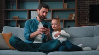 Kafkasyalı baba ve küçük kız çocuğu cep telefonuyla ilgilenen babaya bakıyor çocuk kızına akıllı telefon uygulaması oyunu öğretiyor mutlu aile oyunu oynuyorlar kanepenin üzerinde ebeveynlik eğitimi alıyorlar.