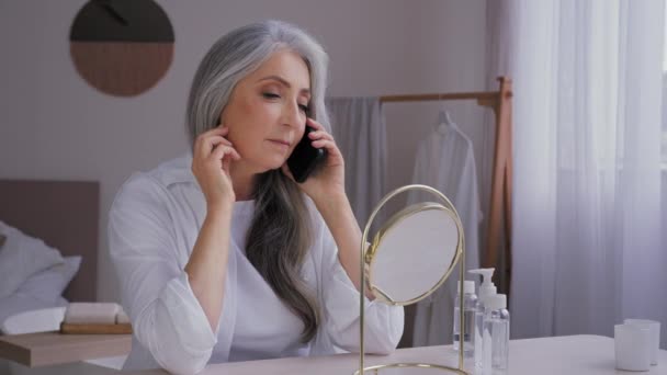 シニア白髪の60代の女性の話携帯電話の話の会話鏡の反射を見て購入するスマートフォンで顔クリームを注文します — ストック動画