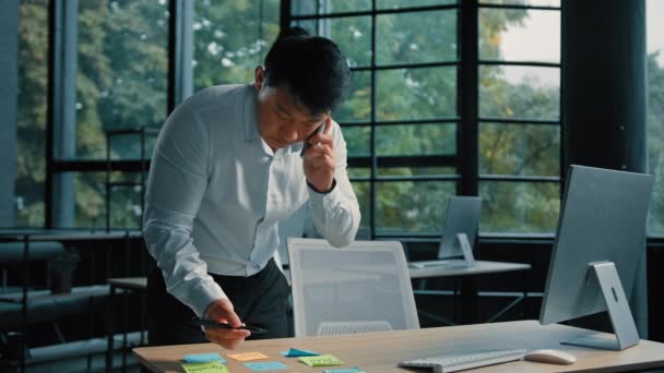 忙しいアジアのビジネスマン韓国人男性が電話で近代的なオフィスの話でテーブルに立って仕事をする携帯電話で専門的な相談を与える粘着ノートプロジェクトの書類の書き込みタスクマルチタスクコンセプト — ストック動画