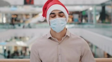 Noel Baba şapkalı adam Covid maskeli adam alışveriş merkezinde dikiliyor kameraya bakıyor hediye kutusunu kırmızı kağıda sarıp Coronavirus salgınını kutluyor.