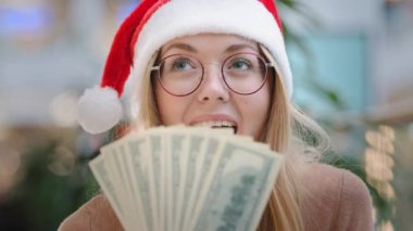 Beyaz kadın Noel Baba şapkası takıyor ve elinde para dolu gözlükler var. Başarılı alışveriş başarısını kutluyor. Ödül ikramiyesi.