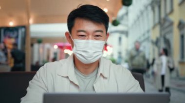 Koruyucu maskeli gülümseyen adam bilgisayarlı ekrandaki sohbete sosyal medyada arkadaşıyla uzaktan kumandalı komik videolar izleyerek e-posta gönderen Asyalı iş adamı turistin internet üzerinden bilgisayar uygulaması kullanarak iletişim kurmasına bak.