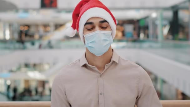 男でサンタクリスマス帽子男男性でCovid Face Maskスタンディングでショッピングセンターショッピングモール見るカメラショーギフトプレゼントクリスマスボックスラップで赤紙お祝いの新年でコロナウイルスパンデミック — ストック動画