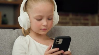 Sevimli beyaz kız çocuk kanepede oturup dinleniyor kulaklıkla müzik dinliyor cep telefonu ekranına bakıyor müzik listesindeki favori pop şarkısının tadını çıkarıyor akıllı telefon cihazıyla müzik konserinde.