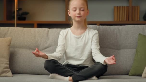 可爱健康的小女孩放松地坐在舒适的沙发上静坐思量家中宁静美丽的少女女儿练习瑜伽呼吸练习闭眼感觉禅和谐 — 图库视频影像