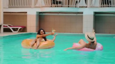 Kafkasyalı Arap kadınlar neşeli, bronzlaşmış kız arkadaşlar lüks spa oteli havuzunda dinleniyorlar yaz tatili şişirilebilir yüzüklerde yüzüyorlar kahkaha kokteylleri içiyorlar.