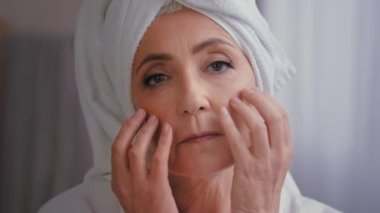 Yaşlı, olgun, 50 'li yaşlarda, kafasında banyo havlusu olan, yüz masajı yapan, buruşuk cilt bakımına dokunan, anti-yaş, nemlendirici krem sıhhatli cildini kaldıran yaşlı, beyaz bir kadın.