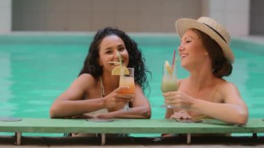 Çekici iki kadın arkadaş Kafkas ve Arap kadın modeller barlarda tropikal alkollü kokteyller içerler yurt dışındaki spa merkezinin havuz kenarında gülerek yaz tatilini kutlarlar.