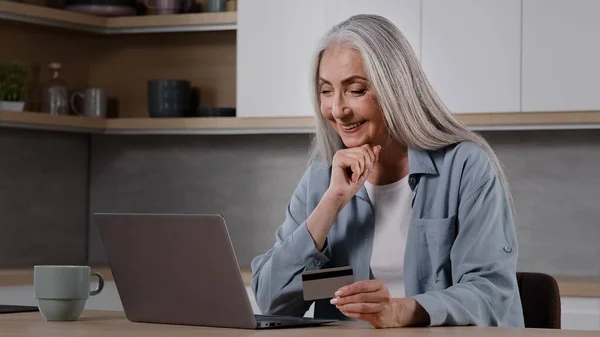 Happy old female caucasian consumer grey-haired granny shopper pay internet order on laptop sits in kitchen hold banking card use online banking app donating money deposit rejoice approved credit loan