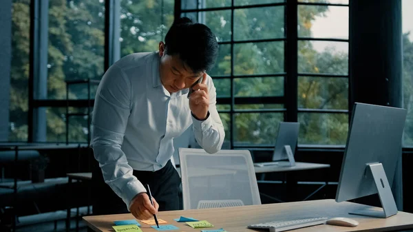 忙しいアジアのビジネスマン韓国人男性が電話で近代的なオフィスの話でテーブルに立って仕事をする携帯電話で専門的な相談を与える粘着ノートプロジェクトの書類の書き込みタスクマルチタスクコンセプト — ストック写真