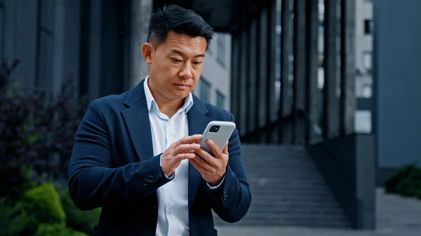 Asian businessman in city has mobile problem with telephone. Middle-aged CEO employer entrepreneur outdoors browsing phone feeling upset with broken mistake error online cellular bad connection lost