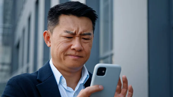 Close up angry worried stressed Asian middle-aged 40s man user businessman employer entrepreneur trying browsing on broken cellphone outdoors in city annoyed with mobile app error connection problem