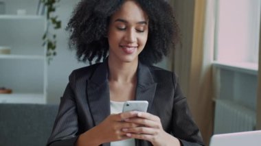 Afrikalı Amerikalı iş kadını serbest çalışan öğrenci kız öğrenci ofiste oturuyor bilgisayarın geliştirdiği uzak eğitim projesiyle cep telefonundan gelen mesajları okuyor modern teknoloji kullanıyor.