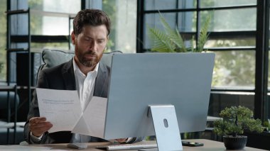 Kafkasyalı dalgın patron müdürü yetişkin 40 'lı yaşlardaki işadamı büroda evrak işleri ve bilgisayar kontrol verileri kağıtlarda arama bilgileri ekrandaki şirket uygulaması projesine bakın