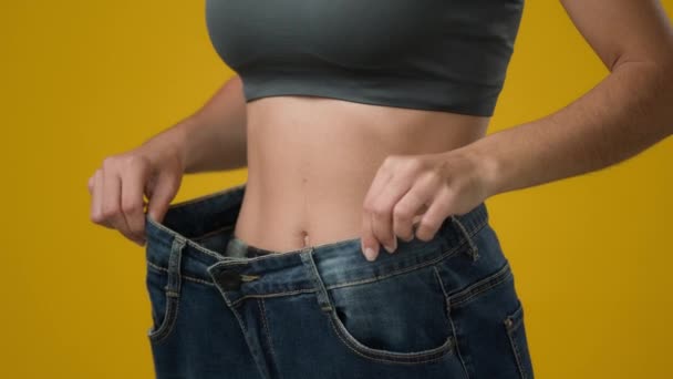 認識できない女の子女性ショー重量損失身に着けている古いジーンズ大きなサイズスリム女性チェック結果の健康的な食生活フィットネススポーツ運動ジム体痩身ウエストでスタジオ黄色の背景 — ストック動画
