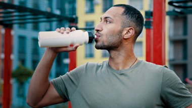 Atletik adam Latin Amerikalı sporcu sıcak içecek soğuk içecek taze su sağlıklı protein içeceği yaz günü susuzluğunu gidermek için şehir spor sahasındaki idmandan sonra dinlendirici konsept konsepti geliştirdi.