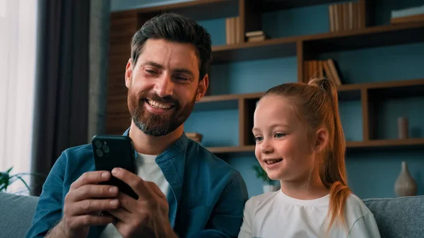 Cute kid preschool girl child looking at smartphone screen with father adult man make selfie photo together use funny face mask app on modern mobile phone gadget laughing watch online video cartoons
