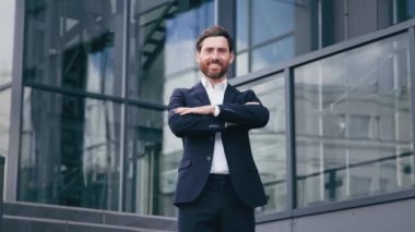 Başarılı, kendine güvenen profesyonel yönetici kollarını kavuşturmuş şirket binasının yanında duran yakışıklı, sakallı, beyaz adam modeli iş merkezinde dışarıda poz veriyor.