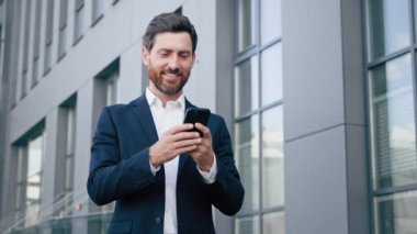 Dışarıda duran mutlu bir adam, elinde telefonla gülümseyen bir işadamı iş için yeni mobil uygulamalar kullanıyor. Onaylama jesti gösteriyor. Mükemmel bir sonuç için en iyi kalite öneriliyor.